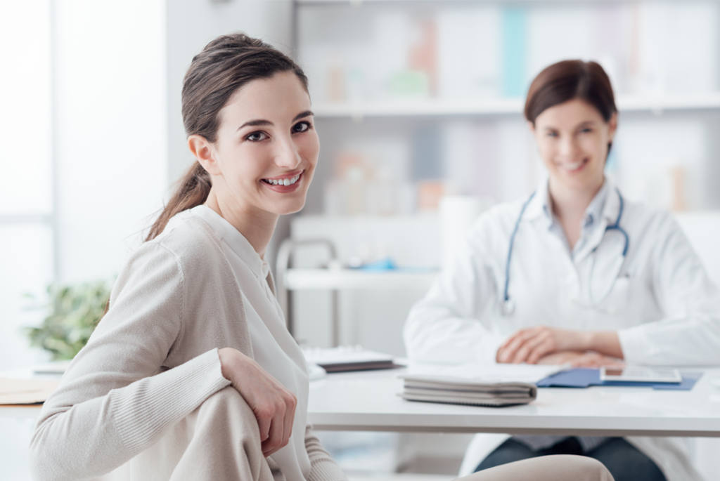 Paciente e médica em consultório. Ambas estão olhando para a foto e sorrindo. A paciente está de costas e virou para a imagem.